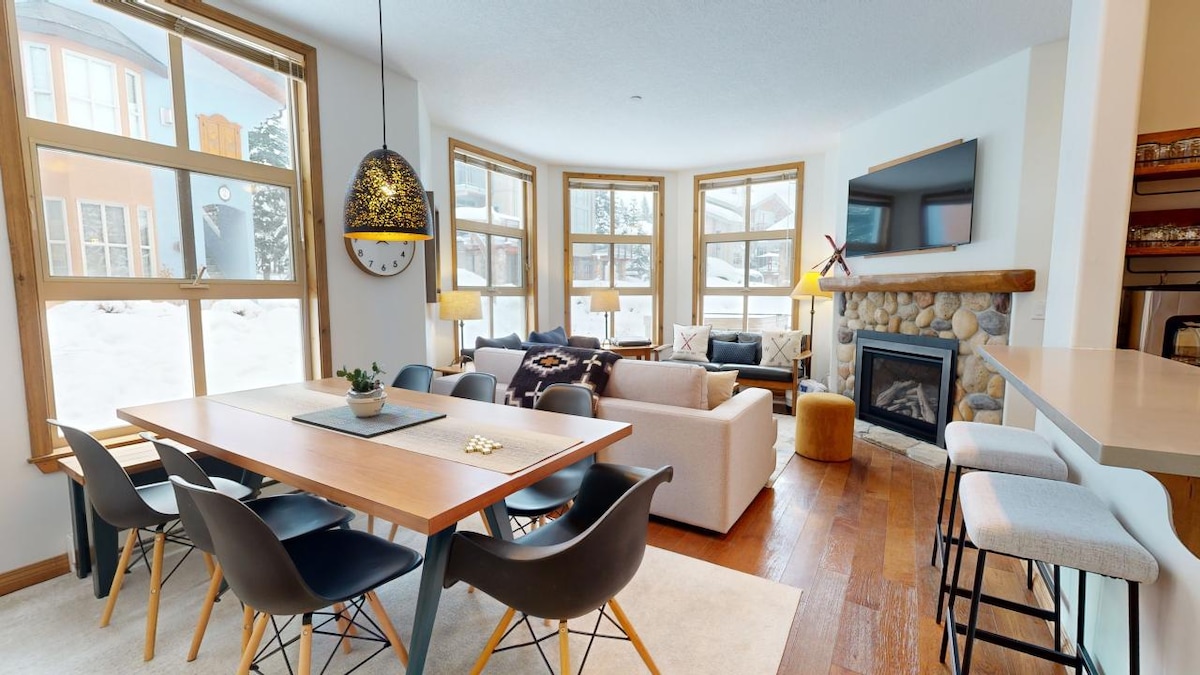 A bright living area is illuminated by large windows, displaying views of the snow-covered landscape. A stone fireplace serves as a focal point, surrounded by comfortable seating. A dining table, paired with modern chairs, is positioned adjacent to the well-equipped kitchen.