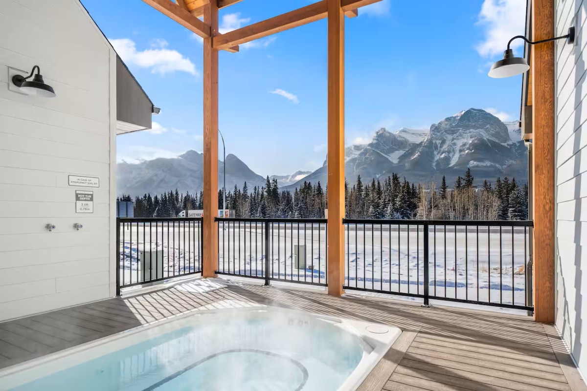 A private patio is shown with a hot tub, framed by wooden beams and a railing. Breathtaking mountain views are visible in the background, and a clear blue sky adds to the serene atmosphere.