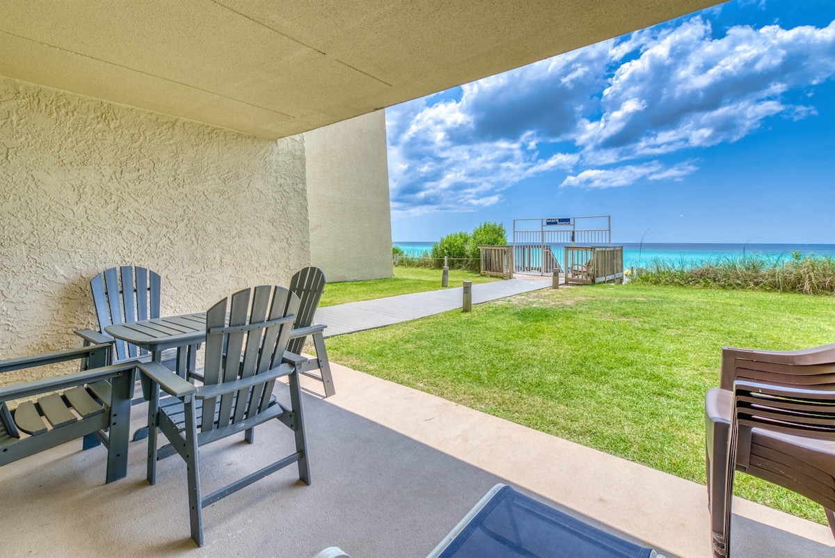 A private beachfront patio showcases comfortable seating with a table and chairs, overlooking a lush green lawn that leads to beach access. The Gulf of Mexico can be seen in the background, framed by a clear blue sky with scattered clouds.
