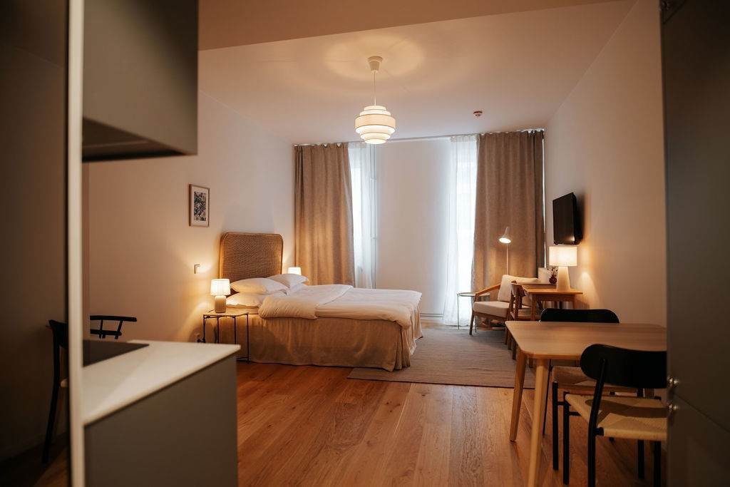 An open-concept studio features a spacious bed with neutral linens positioned against a padded headboard. Natural light filters through sheer curtains. A workspace with a desk and chair is visible, alongside a small dining table with two chairs. A wall-mounted TV adds to the functional decor.