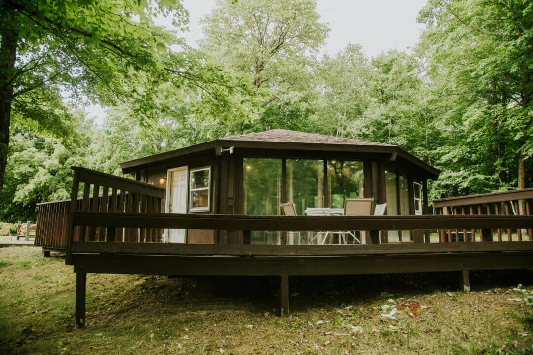 The distinctive octagon cabin is surrounded by lush trees and is complemented by a wraparound deck. The deck features a dining table with multiple chairs, offering an inviting outdoor space. Large windows provide a view of the forest and enhance natural light within.