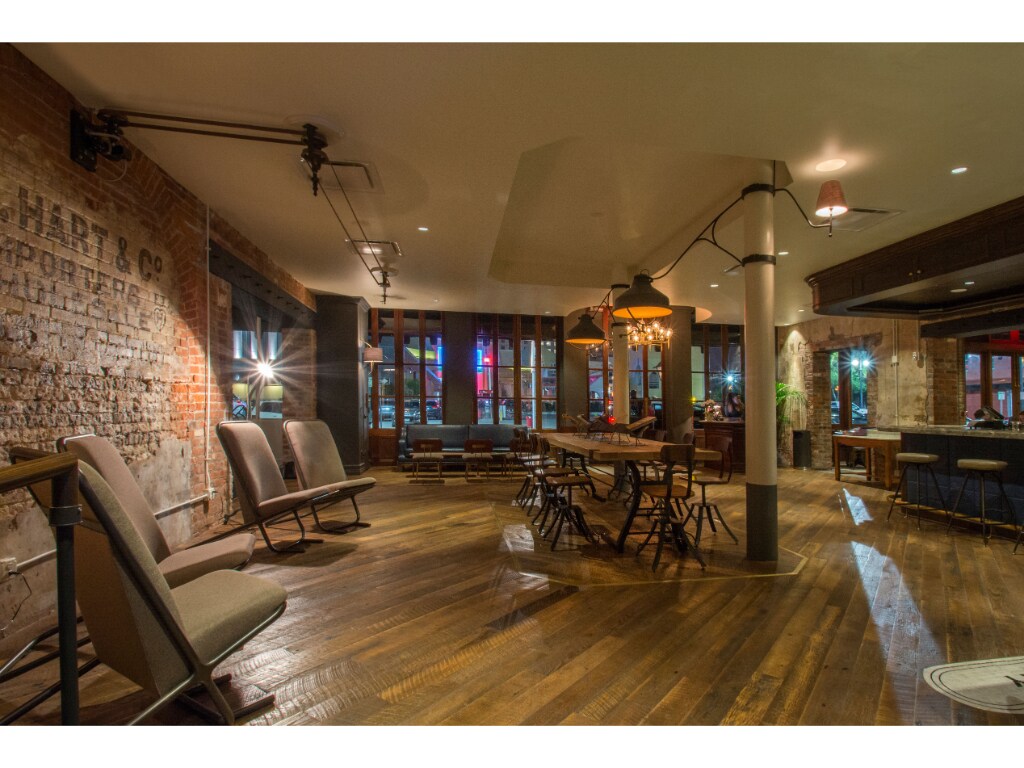 An inviting communal area features exposed brick walls and polished wooden floors. Comfortable seating arrangements include several lounge chairs and a large dining table. Ambient lighting creates a relaxed atmosphere, with windows allowing glimpses of the surroundings.
