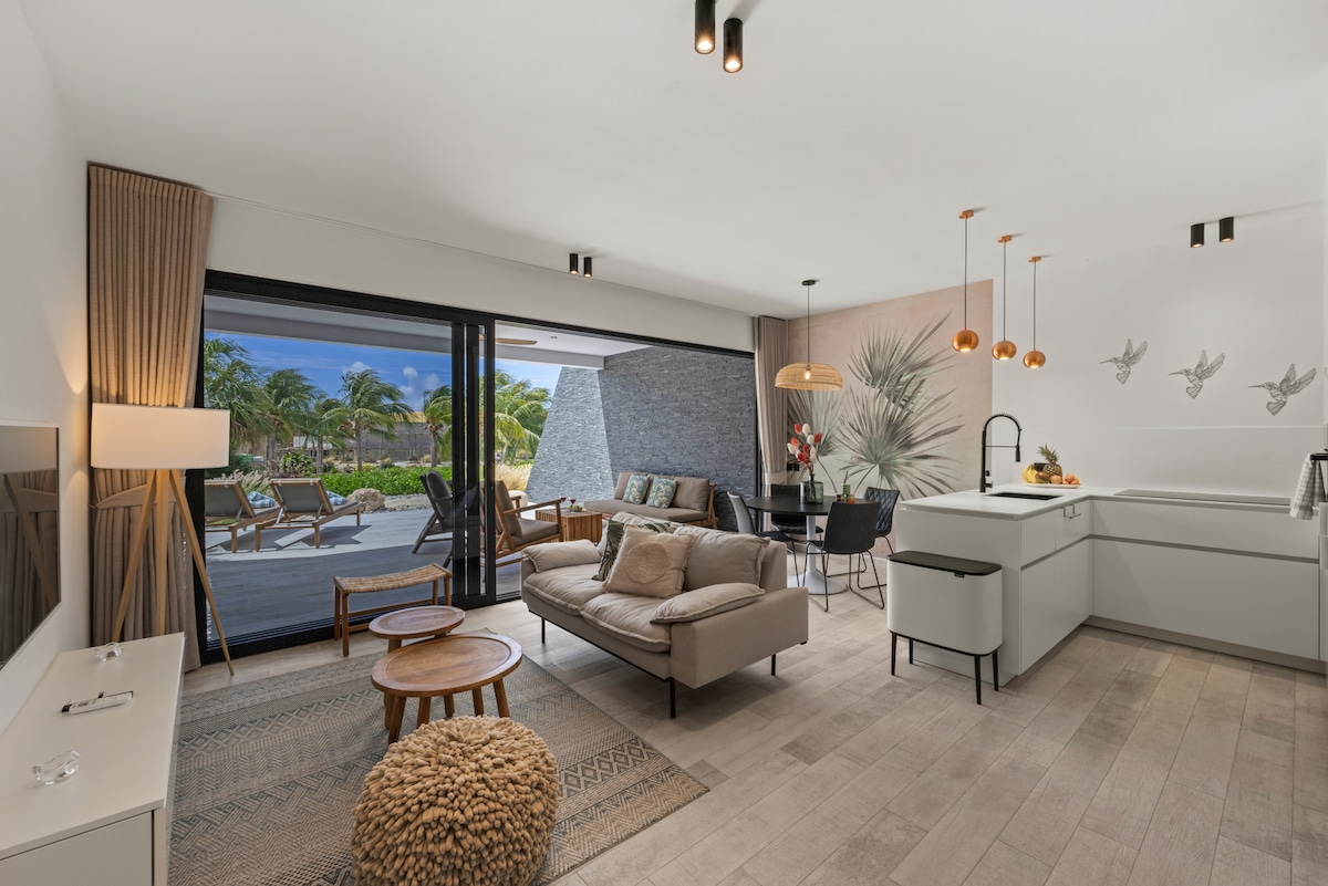 An open-plan living area presents modern decor and sleek furnishings. A comfortable sofa is paired with a textured rug, while natural light floods the space through expansive glass doors that lead to a porch with views of the tropical landscape.