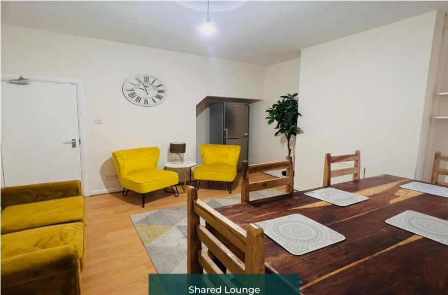 A shared lounge area is depicted, featuring two bright yellow armchairs and a wooden dining table surrounded by chairs. A large clock adorns the wall, and a tall plant adds a touch of greenery. The space is lit with natural light through the window.
