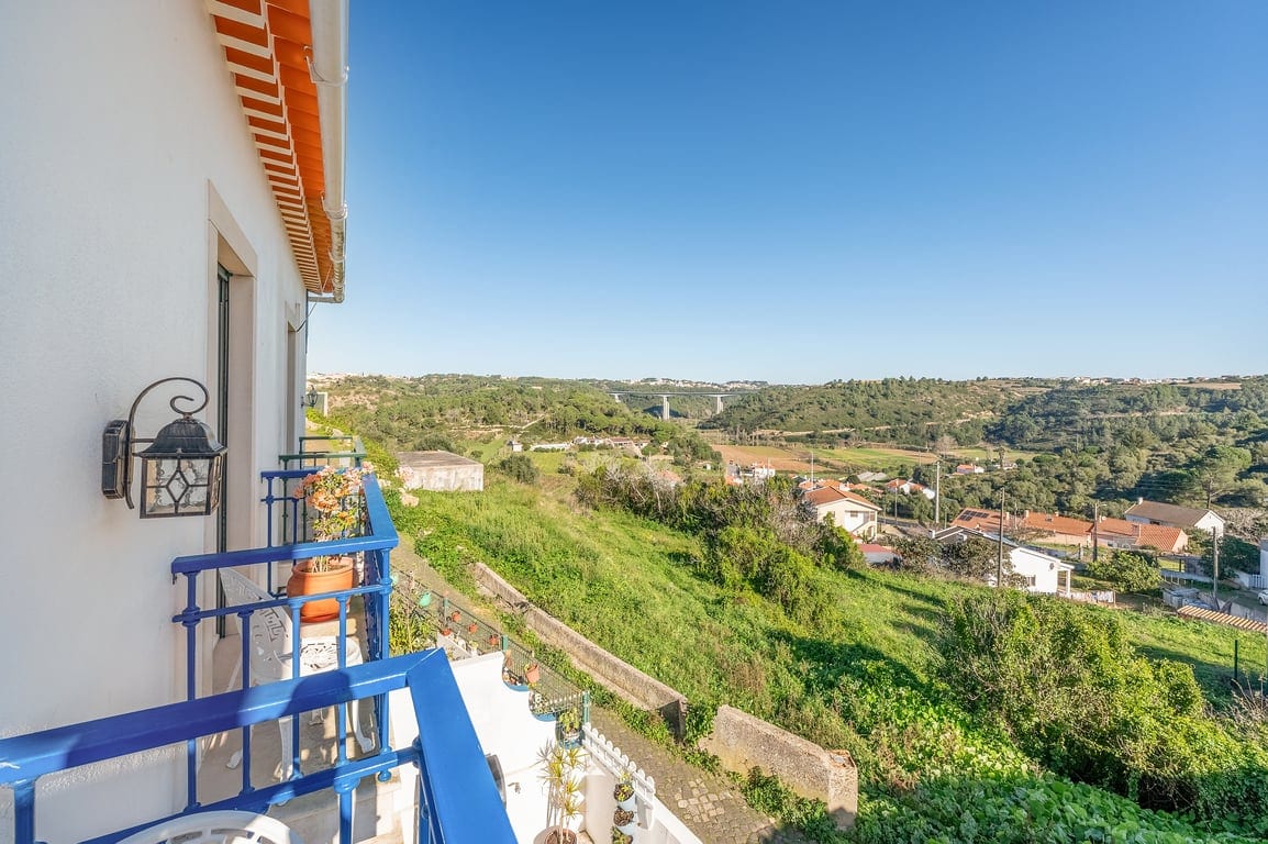 A balcony overlooks a lush green landscape, featuring gentle hills and scattered traditional homes. Blue railings frame the space, while potted plants add a touch of greenery. The clear sky enhances the serene views, providing an inviting outdoor area to enjoy the surroundings.