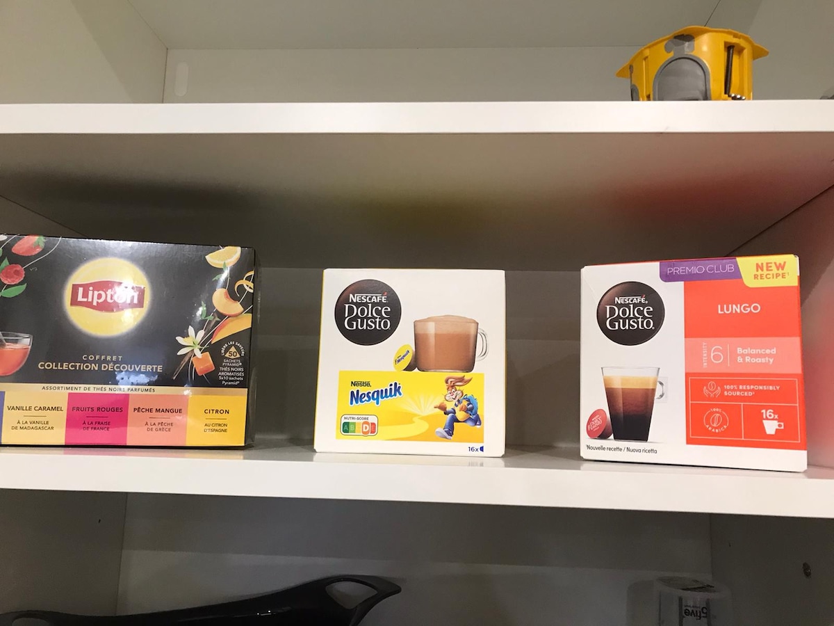 A shelf features three boxes of beverage options. The left box displays Lipton tea assortments, the center box is for Nescafé Dolce Gusto with a Nesquik branding, and the right box offers a new recipe for a coffee blend. A kettle is visible in the background.