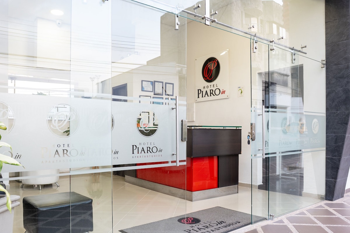 The entrance to Hotel Piaro In is showcased, featuring a glass door with the hotel logo. A sleek reception area is visible beyond the entrance, highlighted by a modern counter and warm lighting. Decorative elements and a welcoming atmosphere invite guests inside.