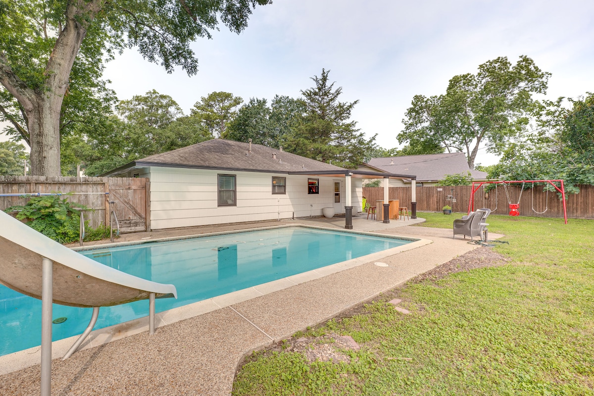 A spacious fenced backyard features an outdoor pool with a slide and a shaded swing set. Comfortable seating is positioned nearby, and a dining area is seen on the patio, surrounded by lush greenery. The inviting pool area is bordered by a textured concrete deck.