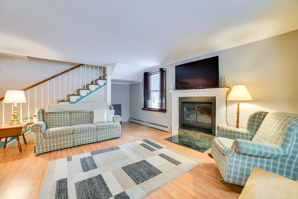 A comfortable living area features two patterned sofas and a light wooden coffee table. A large flat-screen TV is mounted above a decorative fireplace. Natural light illuminates the space, highlighting the warm wood flooring and a patterned area rug that adds texture.