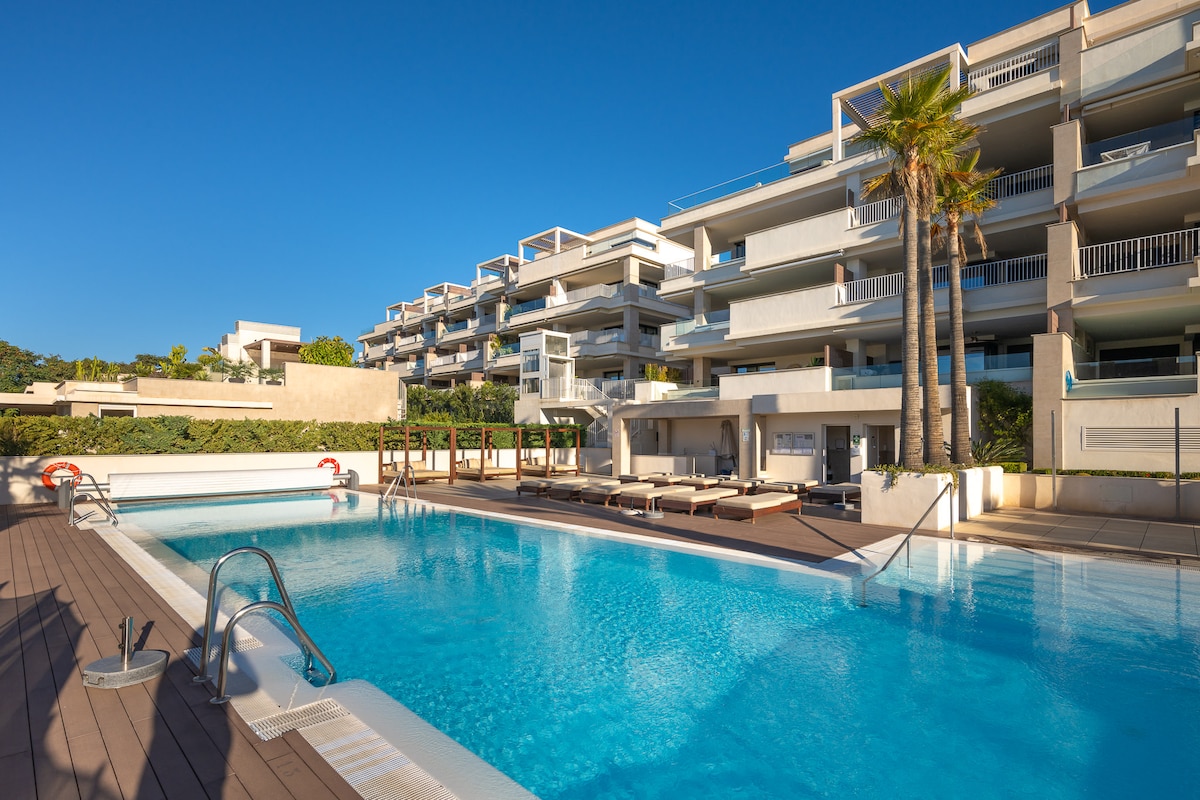 An elegant pool area is showcased, featuring a large swimming pool surrounded by sun loungers. Modern apartments with multiple balconies are visible in the background, enhanced by palm trees. The clear blue sky adds to the serene setting, inviting relaxation and leisure.