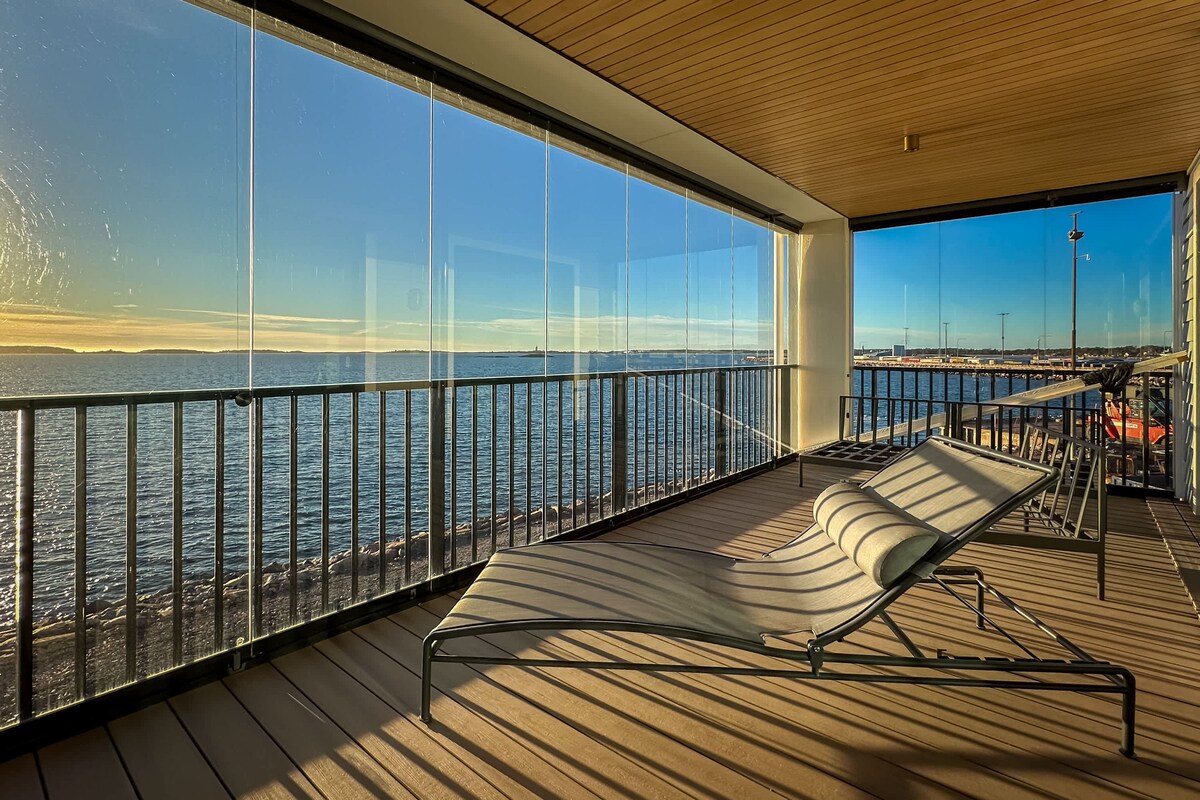 A covered terrace features a chaise lounge positioned for optimal views of the sea. Glass panels provide unobstructed sightlines to the water, while the wooden deck enhances the serene setting, allowing for comfortable outdoor relaxation.