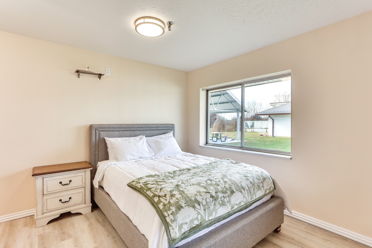 A bright sleeping area features a spacious bed dressed with a white quilt and a green decorative throw. Natural light enters through a large window, offering a view of the outdoor space. A nightstand with two drawers is positioned beside the bed.