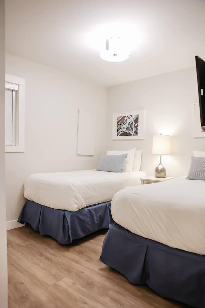 Chumba cha kulala kilicho na vitanda viwili vya mtu mmoja, kinara cha kulala na taa, vyote vikiwa na mwonekano wa upande wowote.