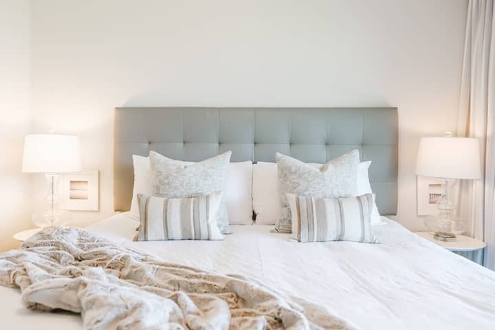 Chumba cha kulala chenye utulivu kilicho na ubao wa kichwa wenye rangi ya kijivu, matandiko meupe na mito yenye mistari, na kuunda mapumziko yenye amani na ya kuvutia.