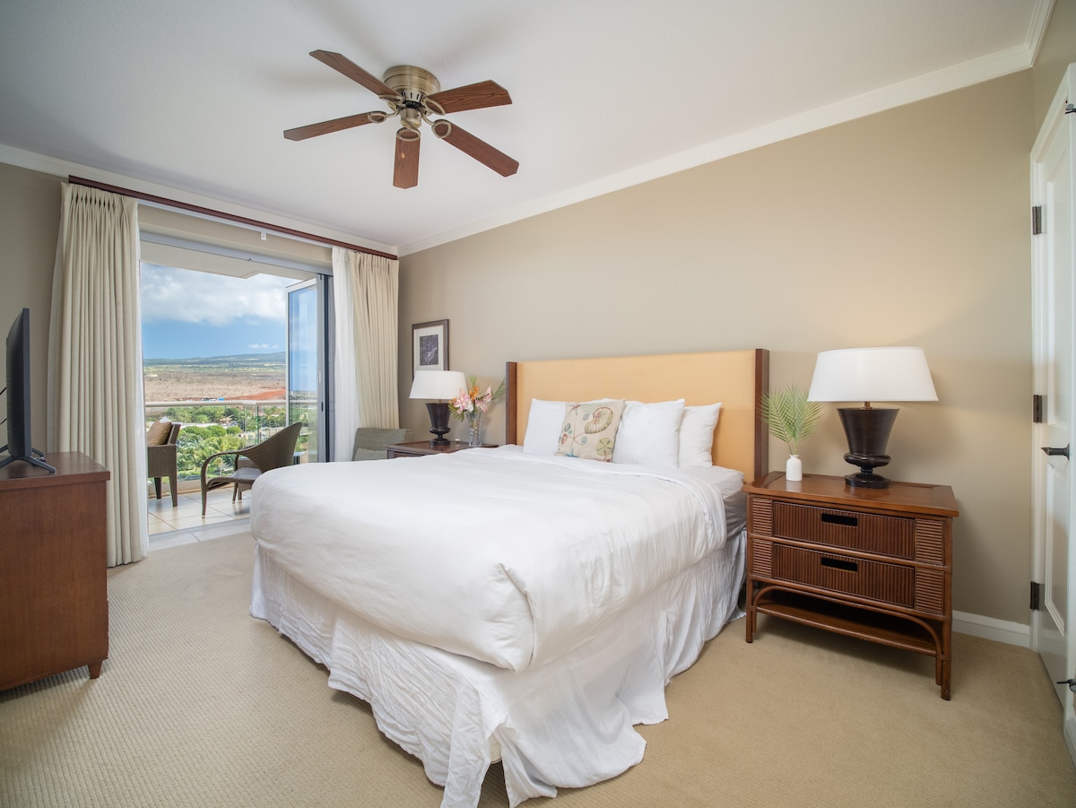 A spacious bedroom features a plush king bed with light bedding, accompanied by two bedside lamps on wicker nightstands. Large windows allow natural light and showcase views of the mountains. A ceiling fan is positioned above, with a cozy seating area visible through the glass doors.