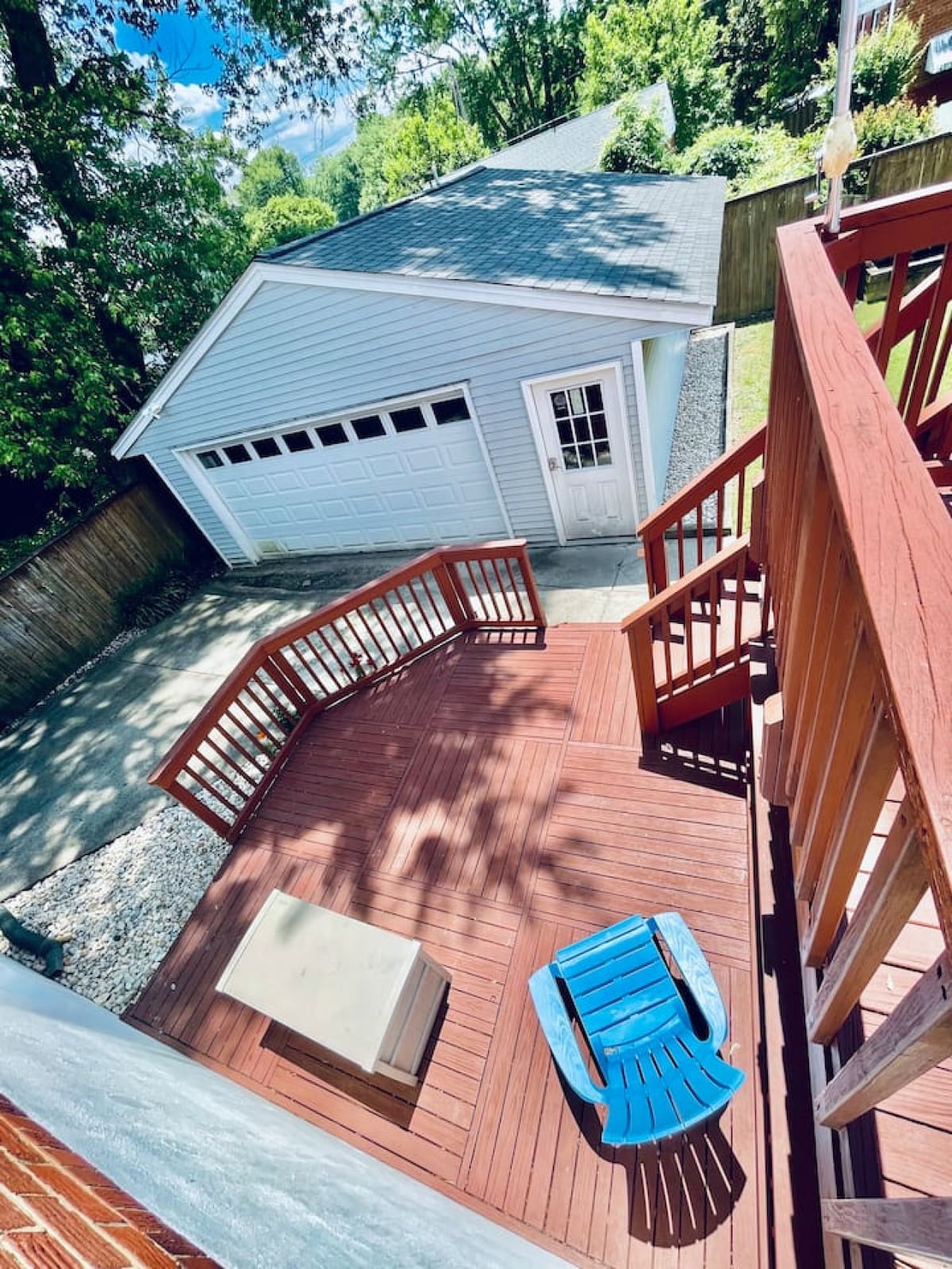 A spacious deck is presented, featuring a blue, single lounge chair positioned near a storage box. A well-maintained garage is visible in the background, along with greenery surrounding the deck area. Sunlight filters through the trees, casting gentle shadows on the deck.