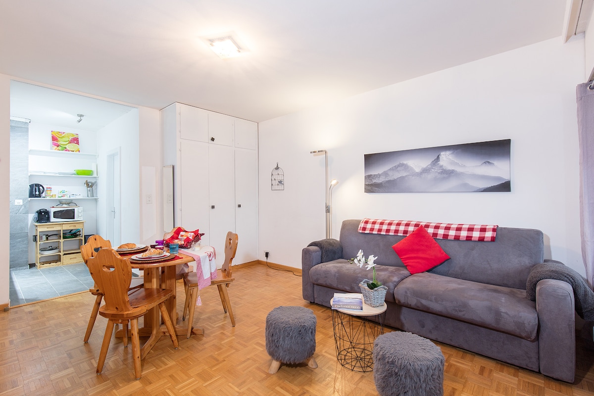 A living area features a comfortable couch adorned with a red accent pillow. A dining table with wooden chairs is set for a meal. A soft rug and poufs create a relaxed atmosphere. The room opens to a glimpse of a modern kitchen area in the background.