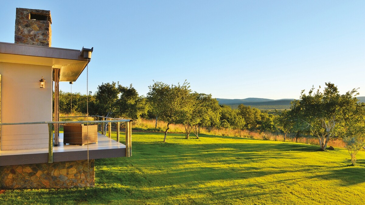 A charming exterior view of the lodge showcases a spacious deck with a wooden railing, leading to a lush green lawn. The landscape features distant hills under a clear blue sky, emphasizing the serene natural surroundings.
