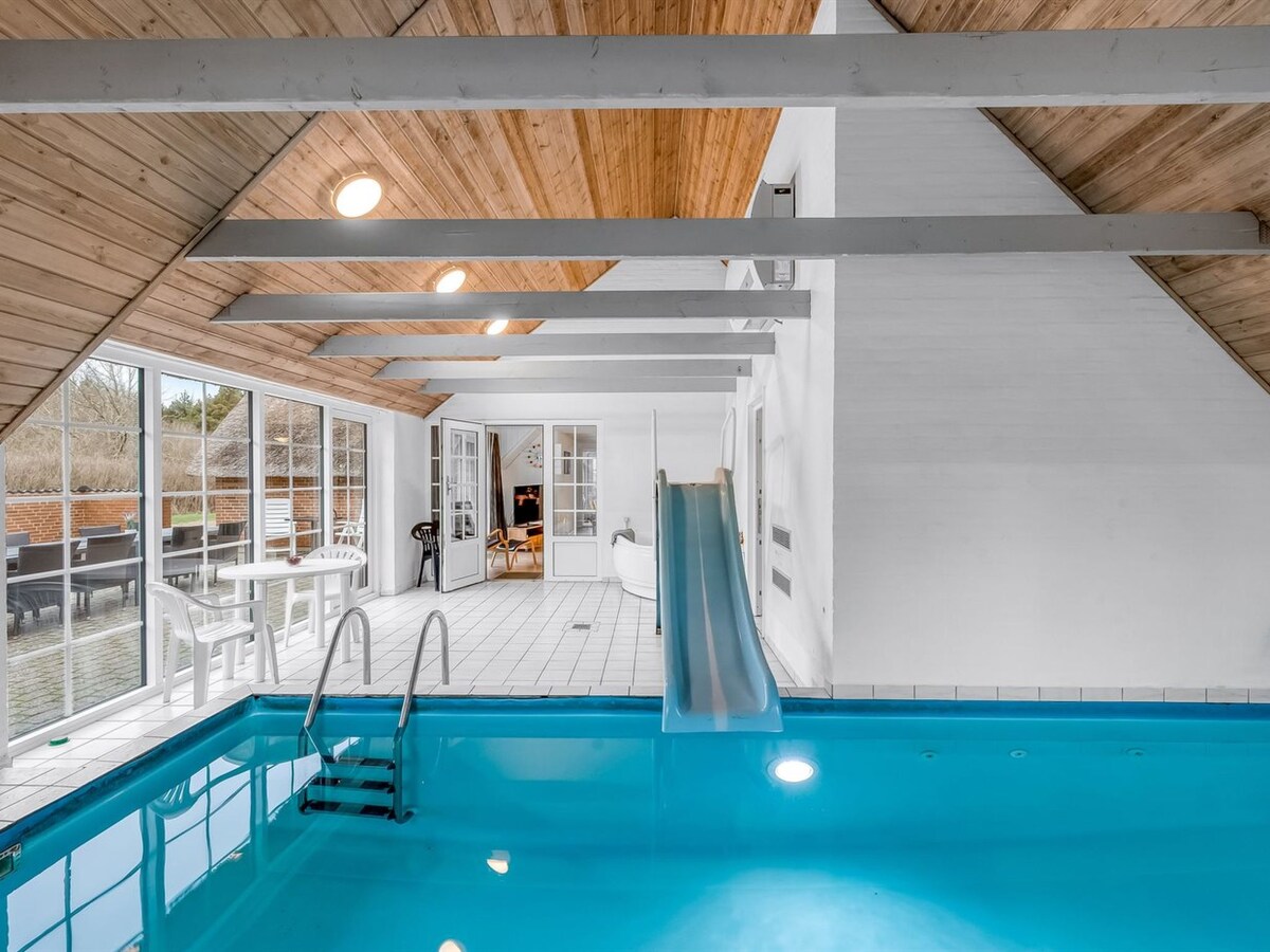 An indoor pool area is displayed, featuring a bright blue pool with a slide. Tall windows allow natural light to fill the space, while wooden beams create an inviting ambiance. Additional seating areas are visible, offering a relaxed environment for recreation.