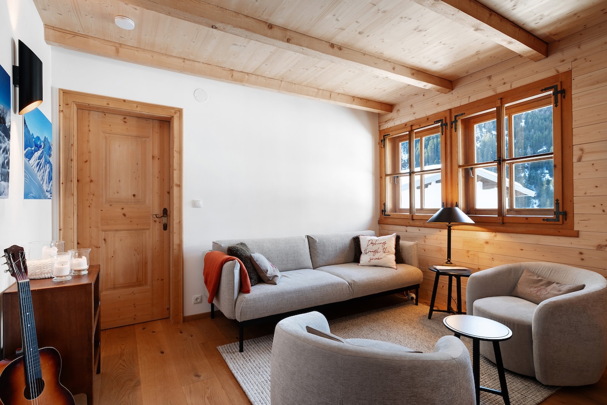 The living room features wooden accents and large windows that allow natural light to illuminate the space. A comfortable gray sofa and two armchairs are complemented by a sleek coffee table. A lamp provides warm lighting, and a small table displays decorative items.