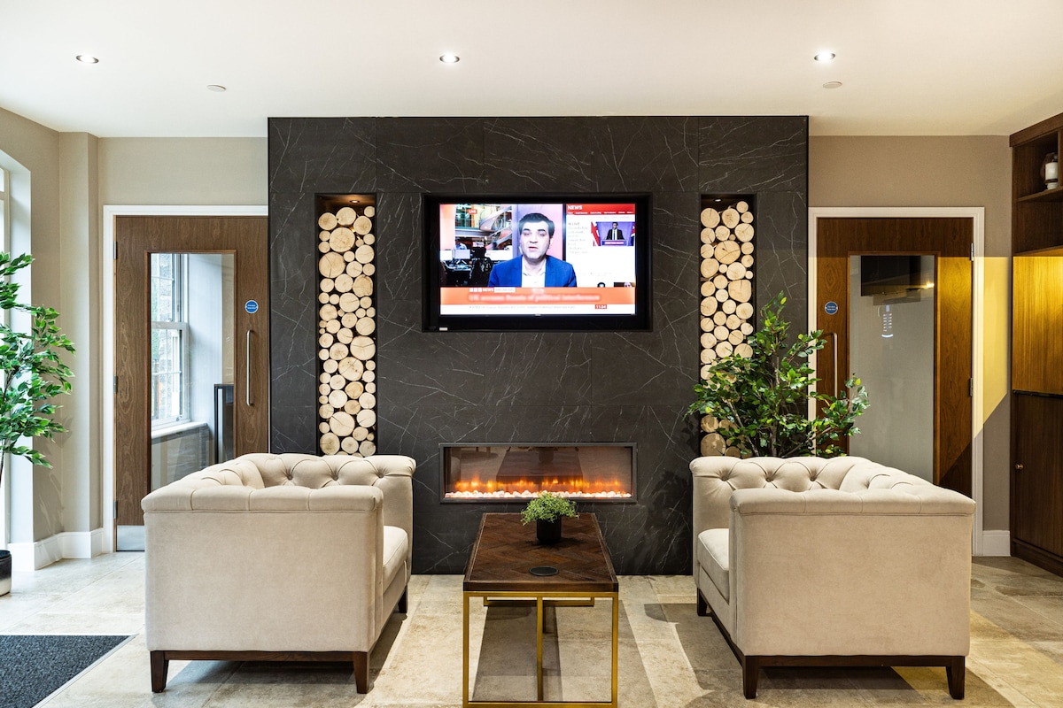 A welcoming lounge area features two plush armchairs facing a low wooden table. An elegant wall with stacked logs acts as a backdrop to a mounted television, providing a cozy setting. Warm lighting highlights the contemporary design and indoor greenery.