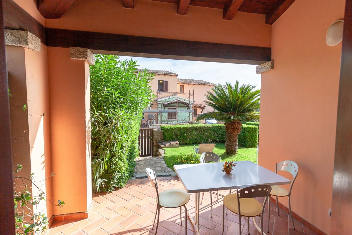 An outdoor veranda features a table surrounded by chairs, overlooking a well-kept garden with lush greenery and palm trees. The entrance is framed by warm-toned walls and wooden beams, providing a calm and inviting space for outdoor dining or relaxation.