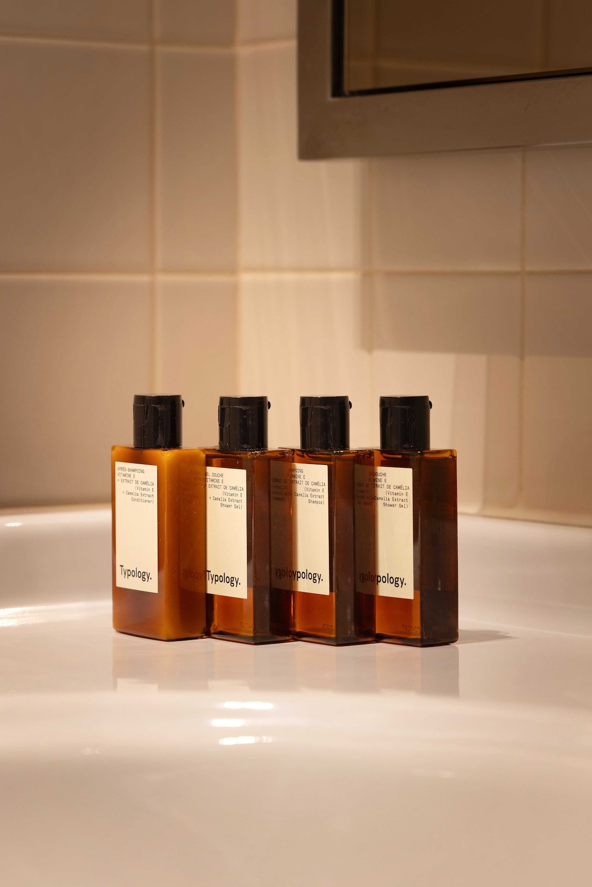 Four bottles of Typology toiletries are neatly arranged on a pristine white sink surface. Each bottle features a simple label, showcasing a cohesive design. The bathroom tiles in the background are light-colored and the overall ambiance appears clean and modern.