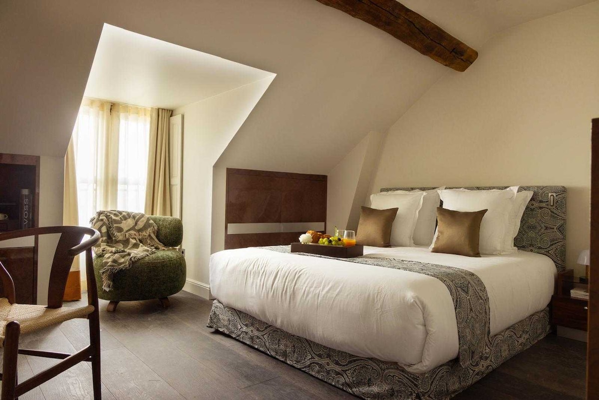 A spacious room hosts a king-size bed adorned with quality linens, complemented by decorative pillows. A cozy seating area is provided with a green armchair. Natural light filters through a large window, accentuating the room's elegant design elements and wooden beam accents.