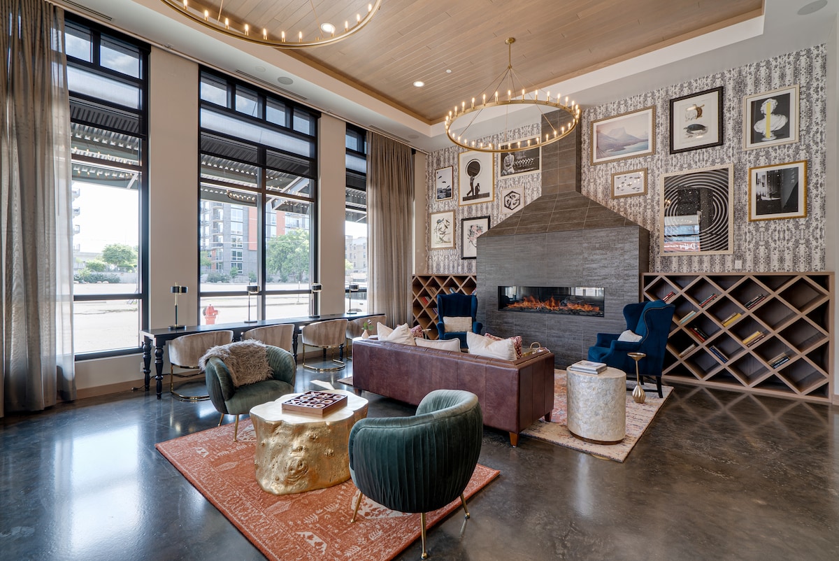 A sophisticated lounge area features large windows that let in natural light, complemented by elegant drapes. Modern seating arrangements include a plush couch and stylish armchairs, while a striking fireplace serves as a focal point. Artwork adorns the walls, adding visual interest to the space.