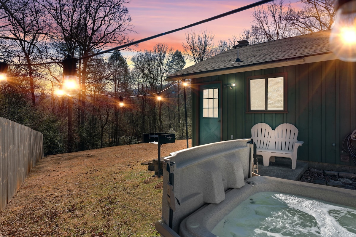 An outdoor hot tub is positioned on a patio, surrounded by a scenic yard. Soft lighting from string lights enhances the inviting atmosphere. In the background, a green cabin and a charcoal grill are visible, framed by trees against a colorful sunset sky.