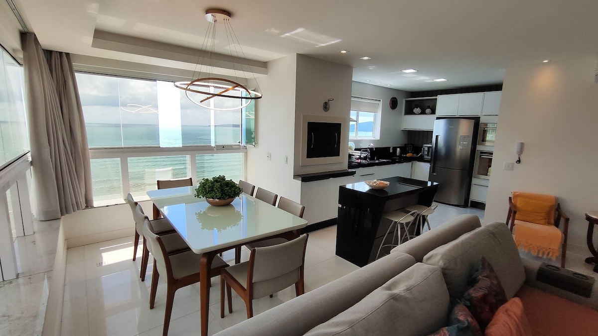 A bright dining area is fitted with a glass table and six chairs, complemented by a cozy living room featuring a sofa and soft armchairs. Large windows reveal stunning sea views, while the modern kitchen, equipped with sleek appliances, is seamlessly integrated into the open space.