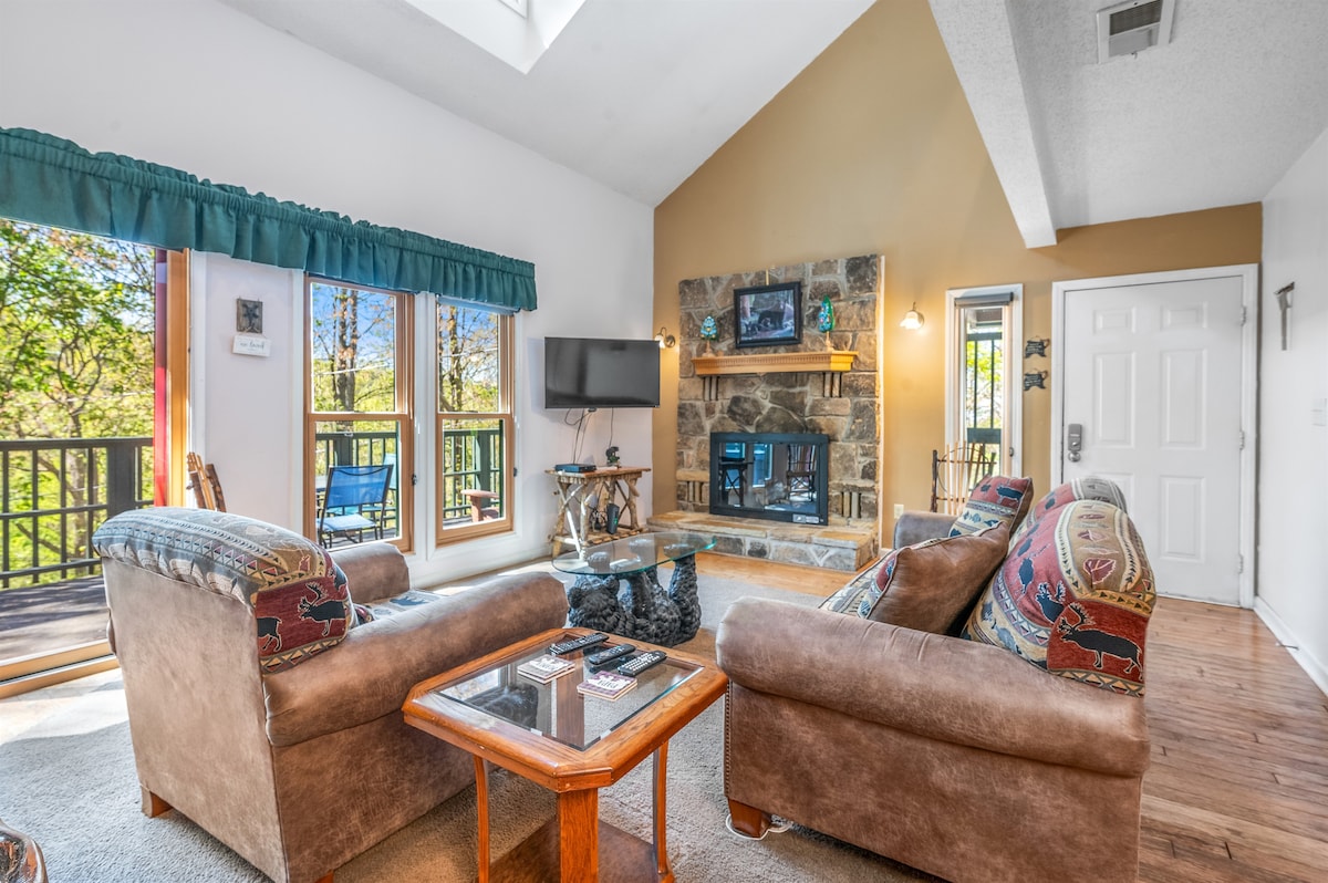 A cozy living room featuring comfortable seating arranged around a coffee table. A stone fireplace is positioned between large windows that provide natural light and views of the outdoors. A flat-screen TV is mounted on the wall, enhancing the inviting atmosphere.