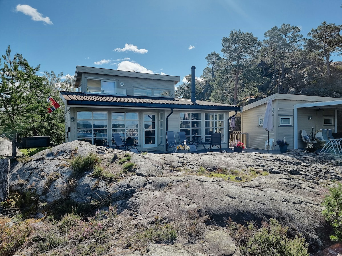 A two-story cottage is positioned on rocky terrain, featuring large windows that allow natural light. Outdoor seating is arranged on a terrace. Surrounding trees offer a sense of privacy. A flag is displayed, adding a touch of local character to the setting.