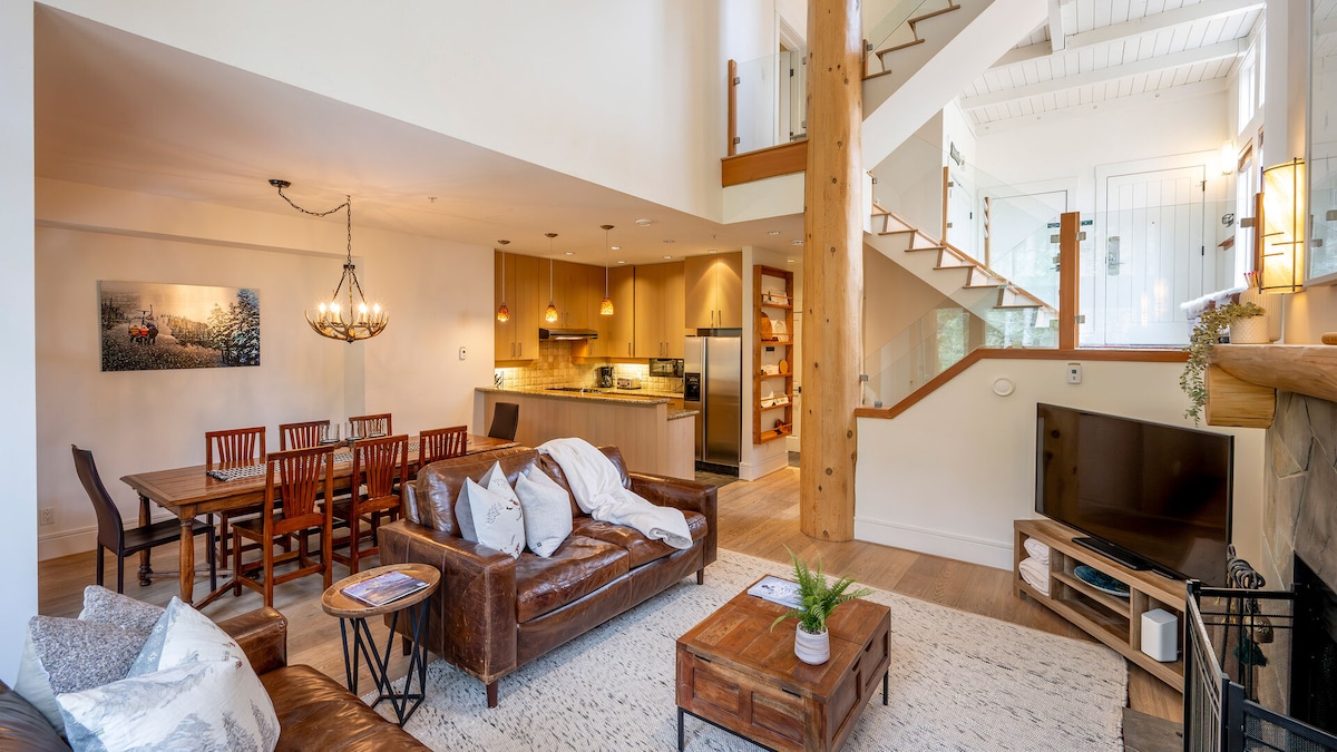 An open living area showcases a leather sofa adorned with blankets, paired with dark wooden furniture including a dining table and chairs. The kitchen features modern appliances and warm wood cabinetry. Large windows create a bright atmosphere, while stairs lead to an upper level.