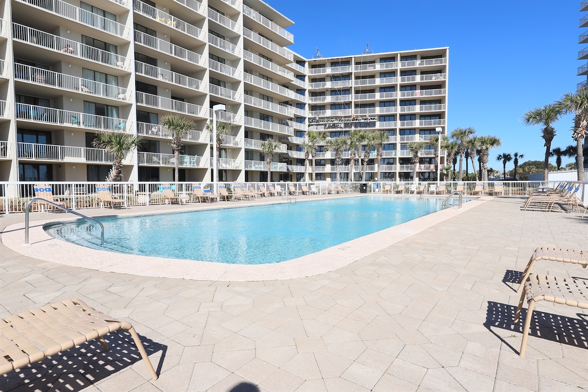 A spacious outdoor pool is surrounded by well-maintained palm trees, with ample seating provided by lounge chairs along the paved area. The height of the surrounding buildings offers a sense of enclosure and privacy, creating a tranquil environment.