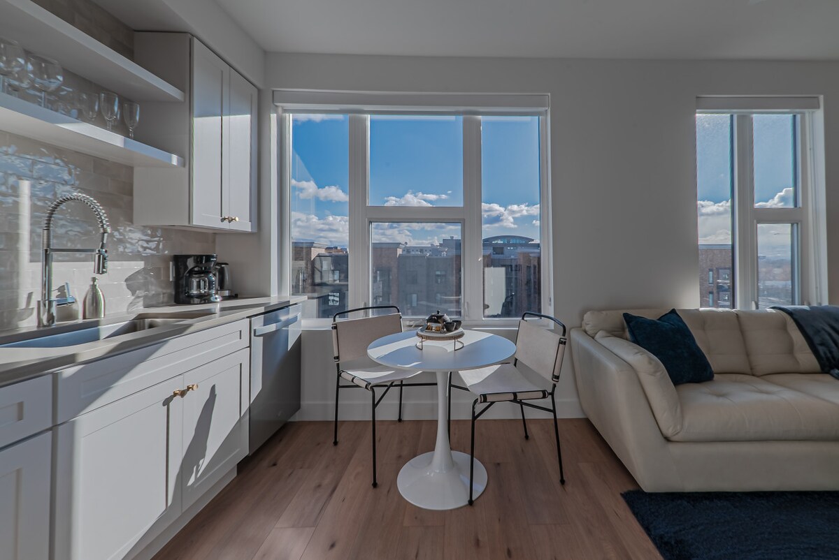 A bright kitchen area features a modern white cabinetry and stainless steel appliances. Natural light floods in through large windows, offering views of the city skyline. A round dining table with two chairs is positioned near the window, complemented by a spacious leather couch.