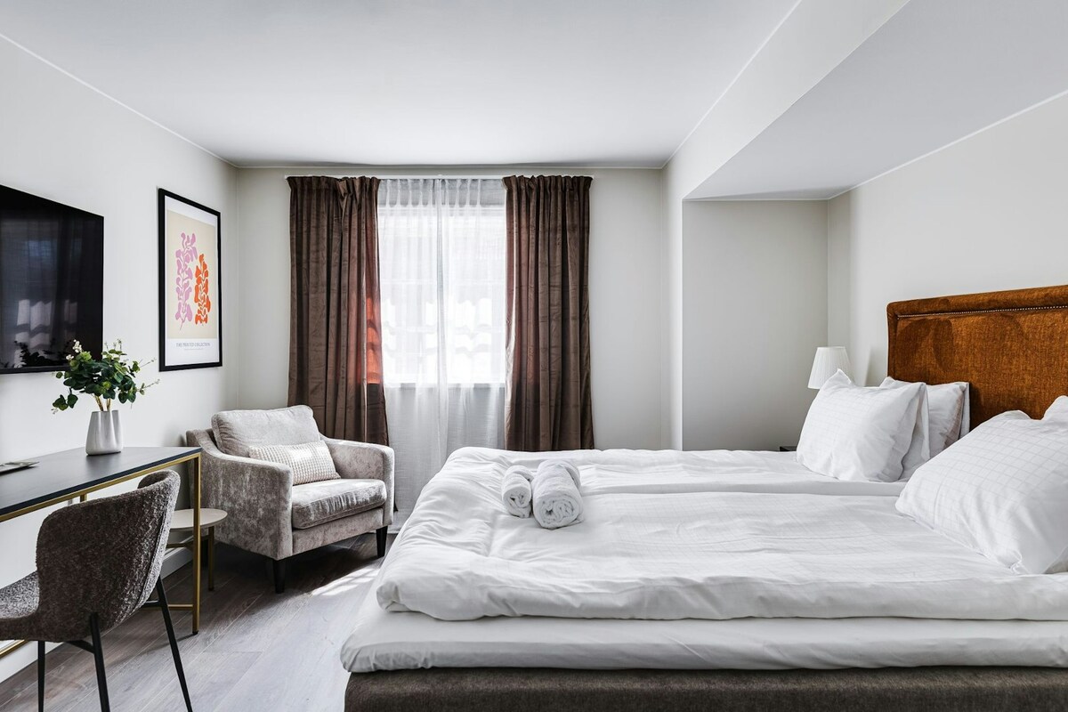 A light and airy bedroom features a spacious bed with crisp white linens. A comfortable armchair is positioned beside a window covered with sheer curtains. A small desk and chair are placed against the wall, with a decorative plant adding a touch of greenery.