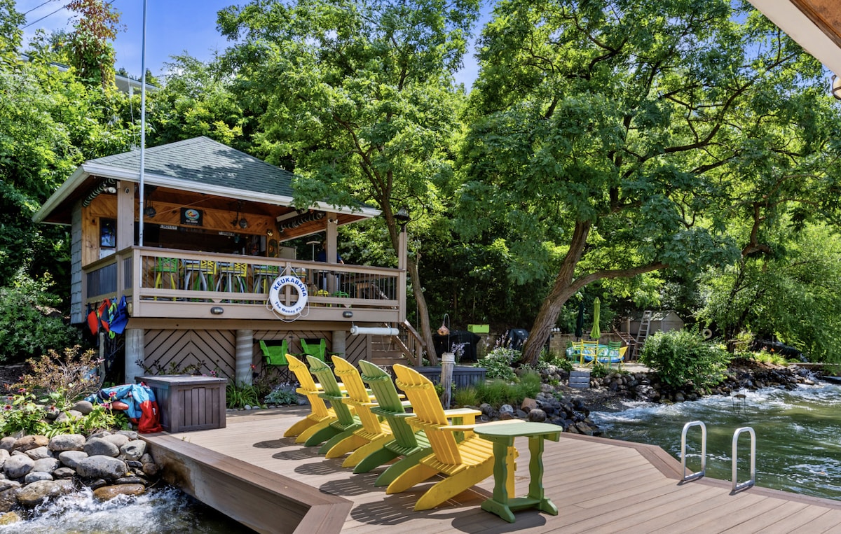 A wooden dock extends towards a serene waterway, featuring four yellow Adirondack chairs arranged for seating. A nearby structure with a green roof houses additional seating and decor. Lush greenery surrounds the area, inviting a sense of tranquility and connection with nature.
