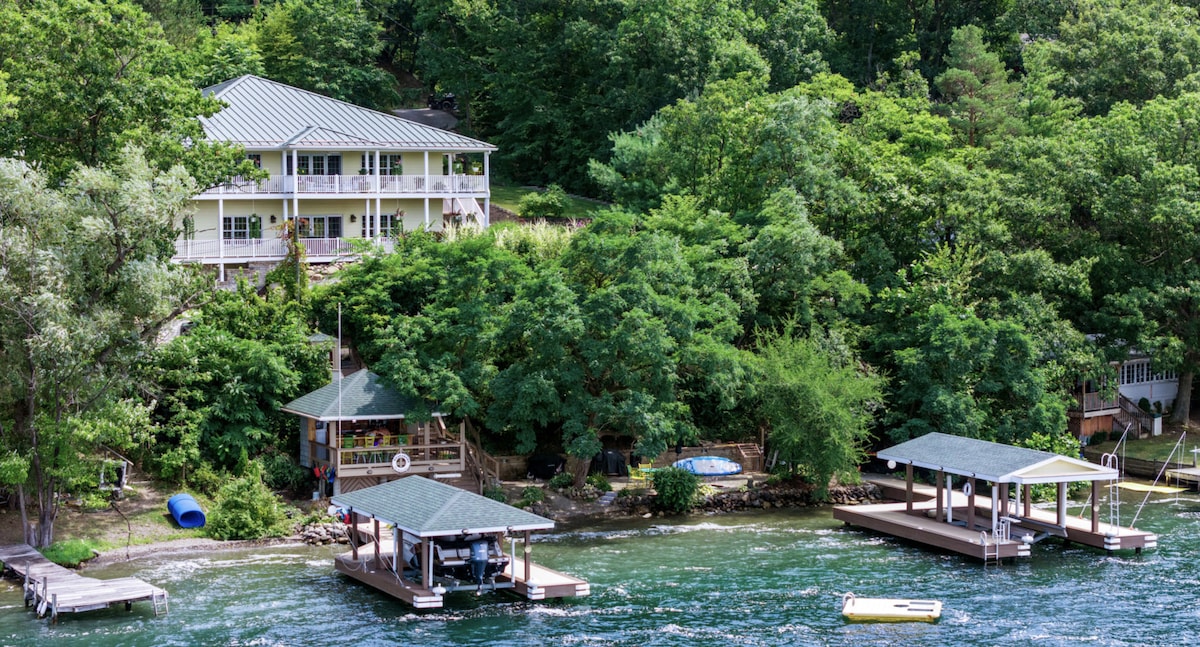 The lakefront lodge is nestled amidst lush greenery, with a two-story structure overlooking the water. Two docks with covered seating areas are visible, alongside a small pool. Kayaks can be seen along the shoreline, contributing to the serene lakeside environment.