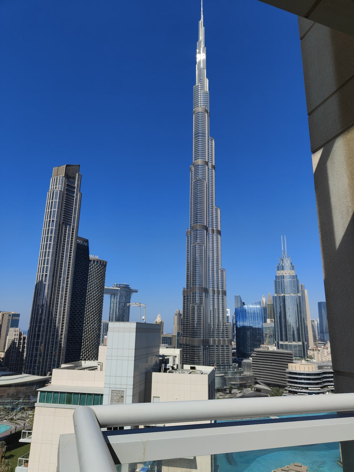 The image features a stunning view of the Burj Khalifa, towering prominently against a clear blue sky. Surrounding buildings are visible, showcasing Dubai's modern skyline, with a hint of greenery from the nearby structures in the foreground.