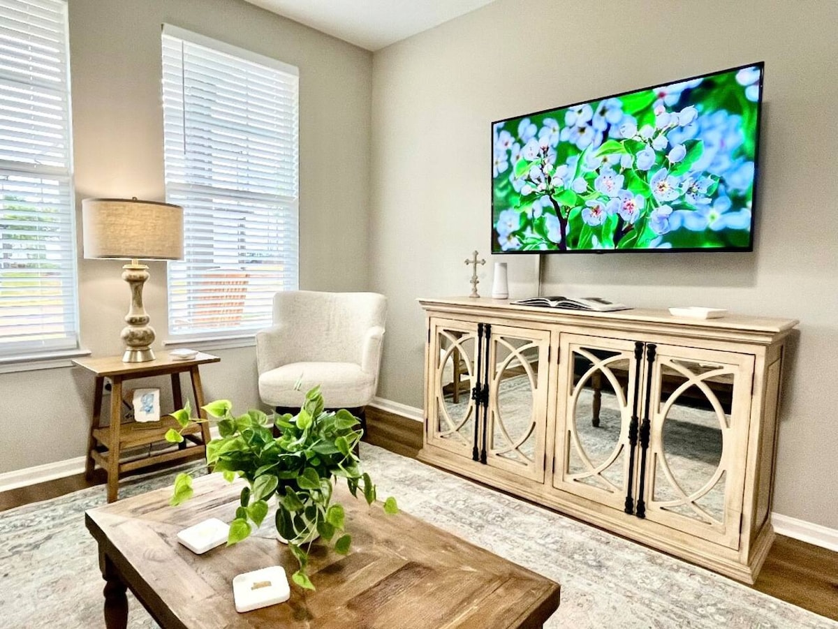 A bright living area features a large flat-screen television displaying floral imagery. A light-colored armchair and wooden side table accompany a decorative console table, which showcases dark accents. Natural light floods in through two windows adorned with light blinds, enhancing the neutral tones of the space.