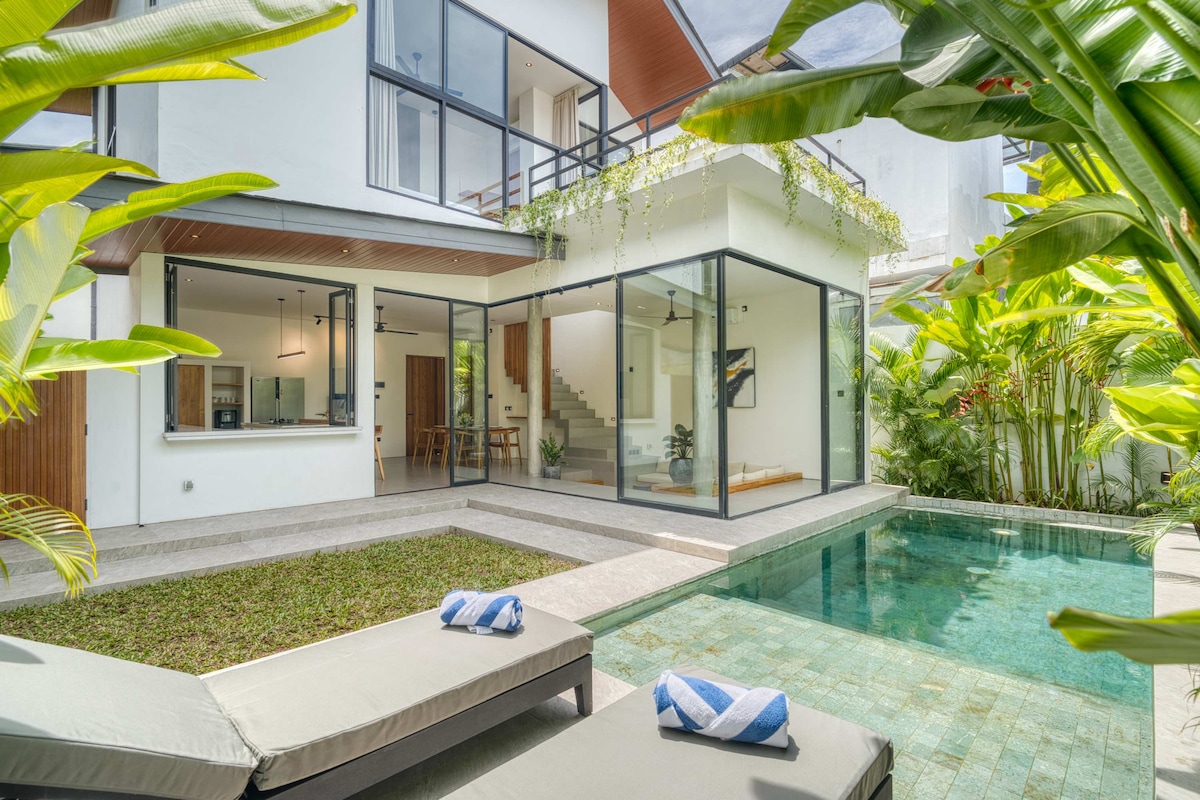 The villa's outdoor area features a crystal-clear swimming pool surrounded by greenery, with comfortable sunbeds positioned beside it. Large glass doors provide a view into a bright living space, while tropical plants create a lush backdrop.