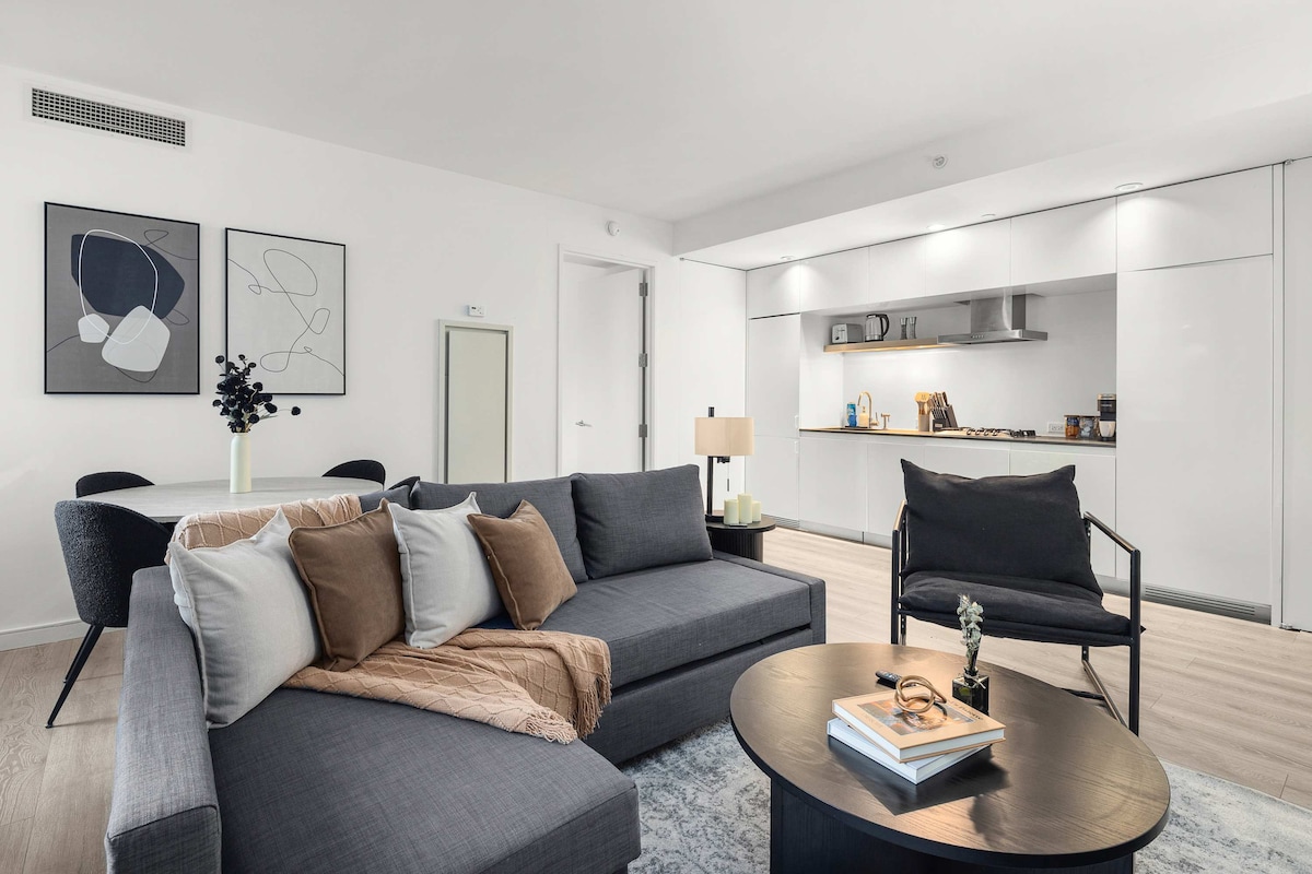 A modern living area features a gray sectional sofa adorned with neutral and pastel cushions. A round coffee table is situated in front, displaying books and decor. The open-plan kitchen is visible in the background, equipped with sleek appliances and cabinetry.