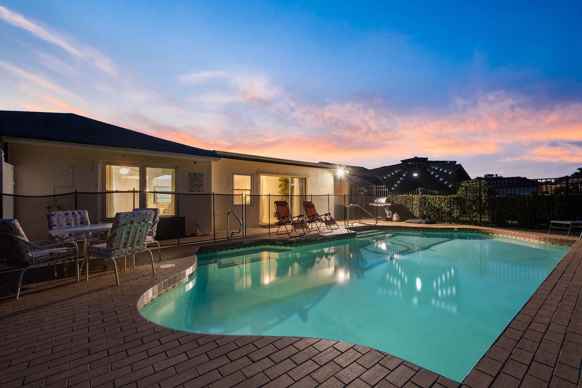 The serene swimming pool is surrounded by a paved patio, reflecting the vibrant hues of the sunset sky. Lounge chairs are positioned nearby, inviting relaxation. The spacious home features large windows, allowing for seamless indoor-outdoor access and showcasing the peaceful ambiance.