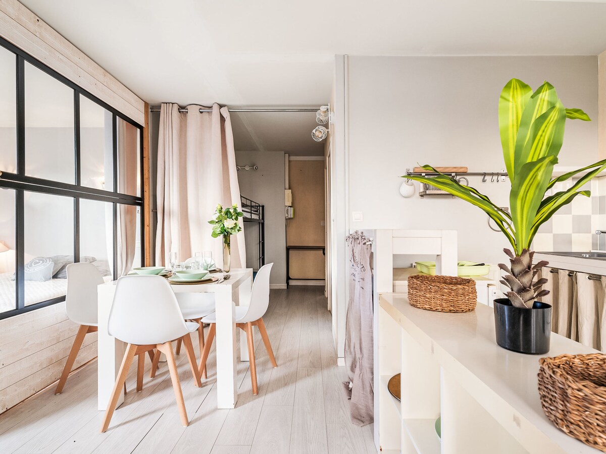 The dining area features a light wooden table surrounded by white chairs, set for a meal. A potted plant adds a touch of greenery. Natural light is allowed in through large windows, with sheer curtains providing privacy. The space is connected to an open kitchen area.