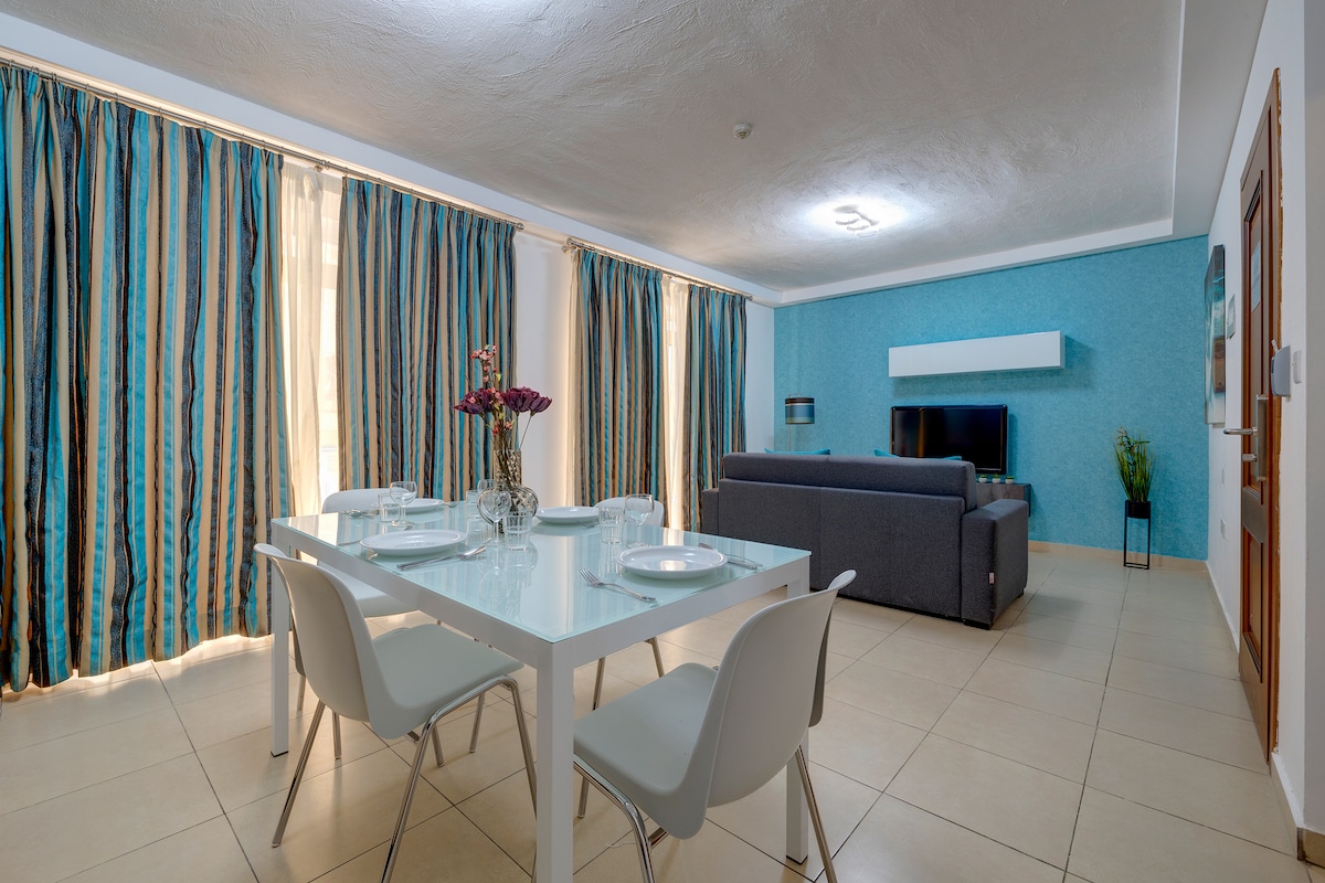 A modern living area features a comfortable gray sofa facing a wall-mounted television. A white dining table is set for four, accompanied by sleek, white chairs. Light pours in through large windows, framed by blue and gold striped curtains.