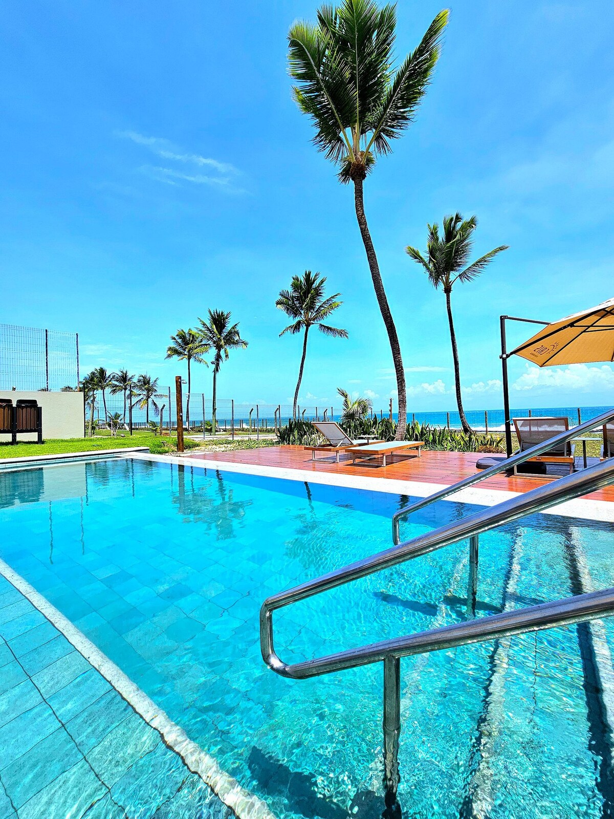 A clear swimming pool reflects the blue sky, with palm trees swaying alongside. Sun loungers are placed on a wooden deck, inviting relaxation. The serene water appears inviting, and a section of green landscape is displayed in the background.