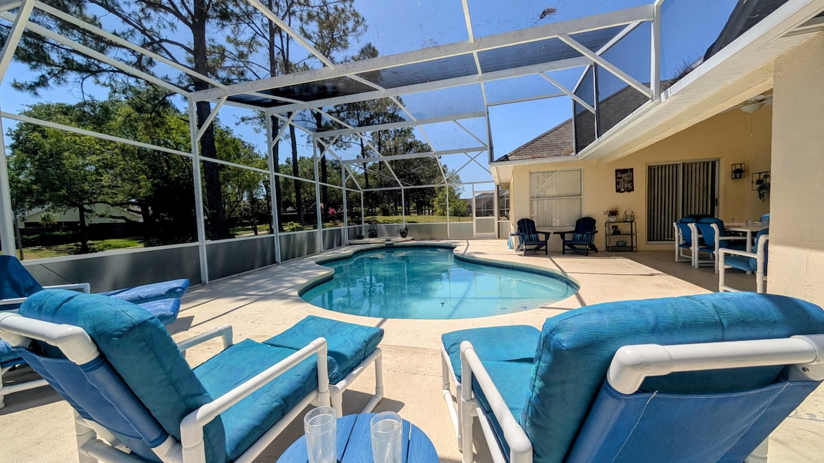 The pool area features a spacious deck with a clear enclosure providing a view of the surrounding greenery. Light blue lounge chairs are arranged around the pool, which is shaped organically. Sunlight reflects off the water's surface, creating a serene atmosphere.