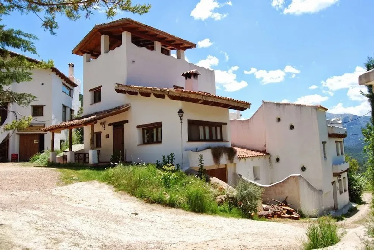 A spacious apartment complex is set against a backdrop of clear blue skies and lush greenery. The white exterior is complemented by rustic wooden details, and a unique roof peak adds charm. The area is surrounded by well-tended landscaping, enhancing the inviting atmosphere.