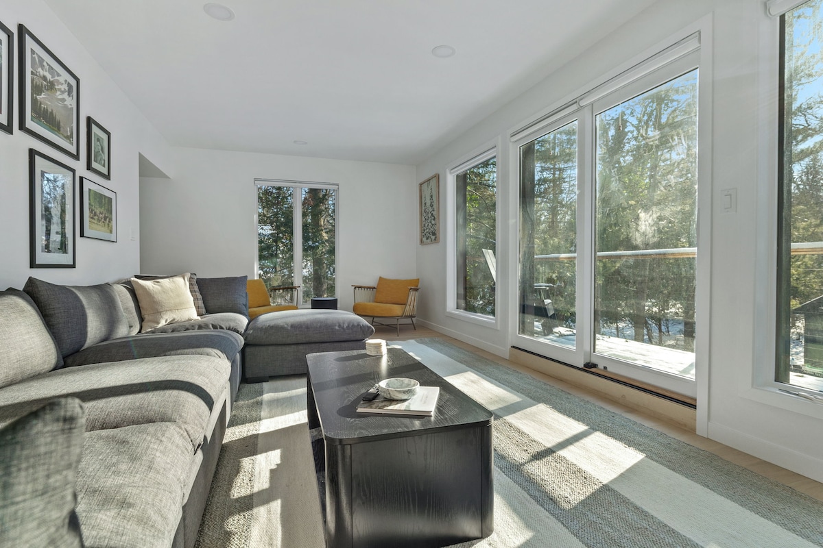 A modern living area is showcased with a spacious sectional sofa and a sleek coffee table. Floor-to-ceiling windows invite natural light, revealing views of the surrounding trees. Subtle wall art adds character, while a cozy accent chair enhances the welcoming ambiance.